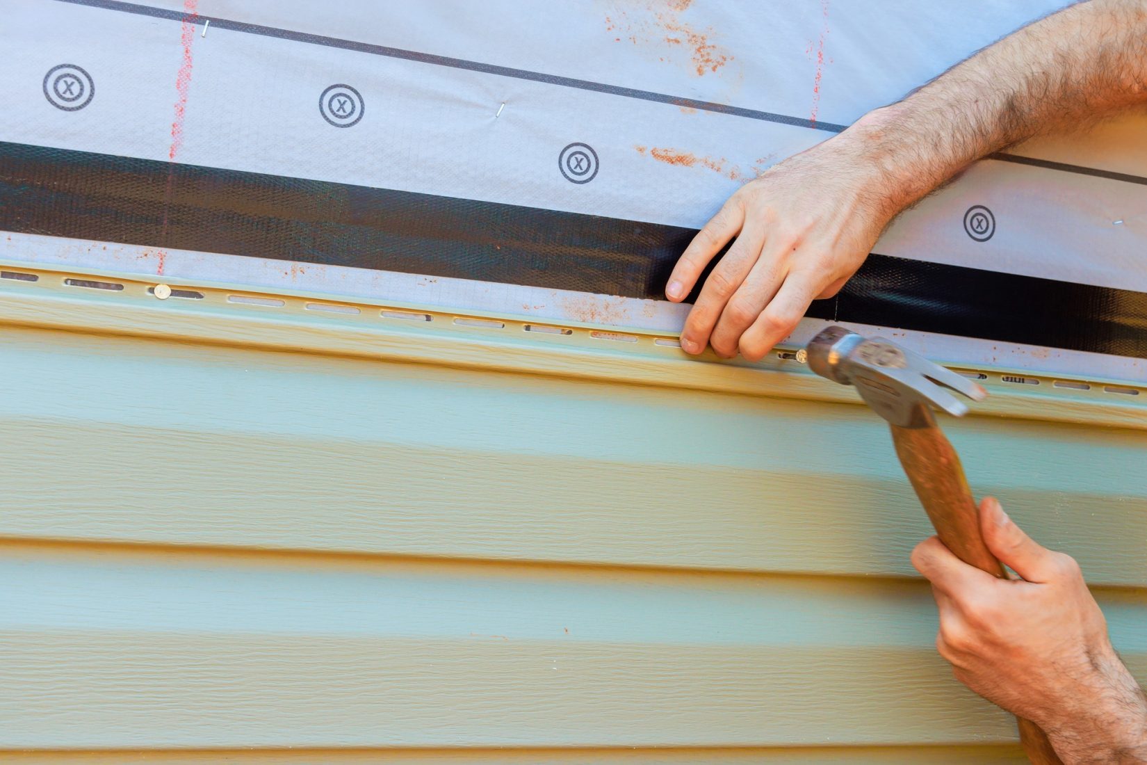 Hands installing house siding with hammer and protective materials